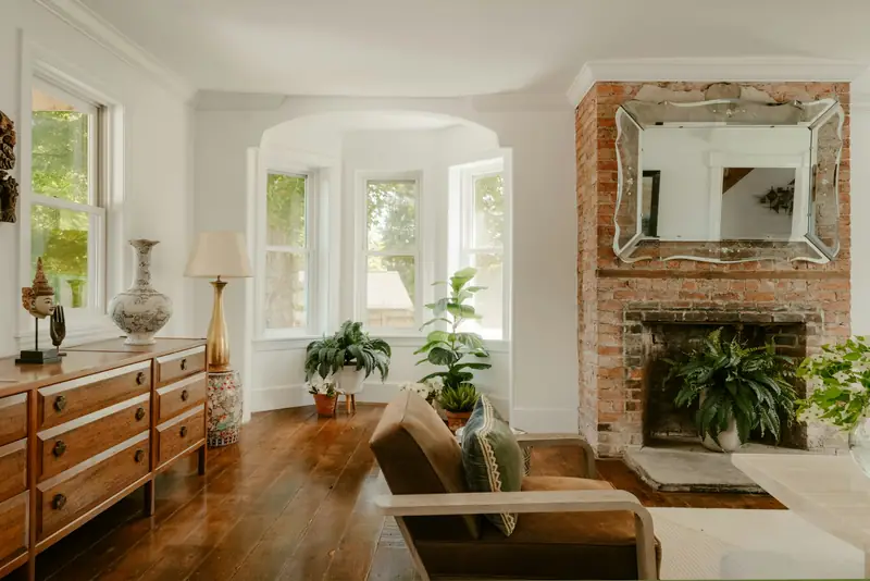 A brick fireplace with a Venetian mirror and a walnut dresser by a bay window